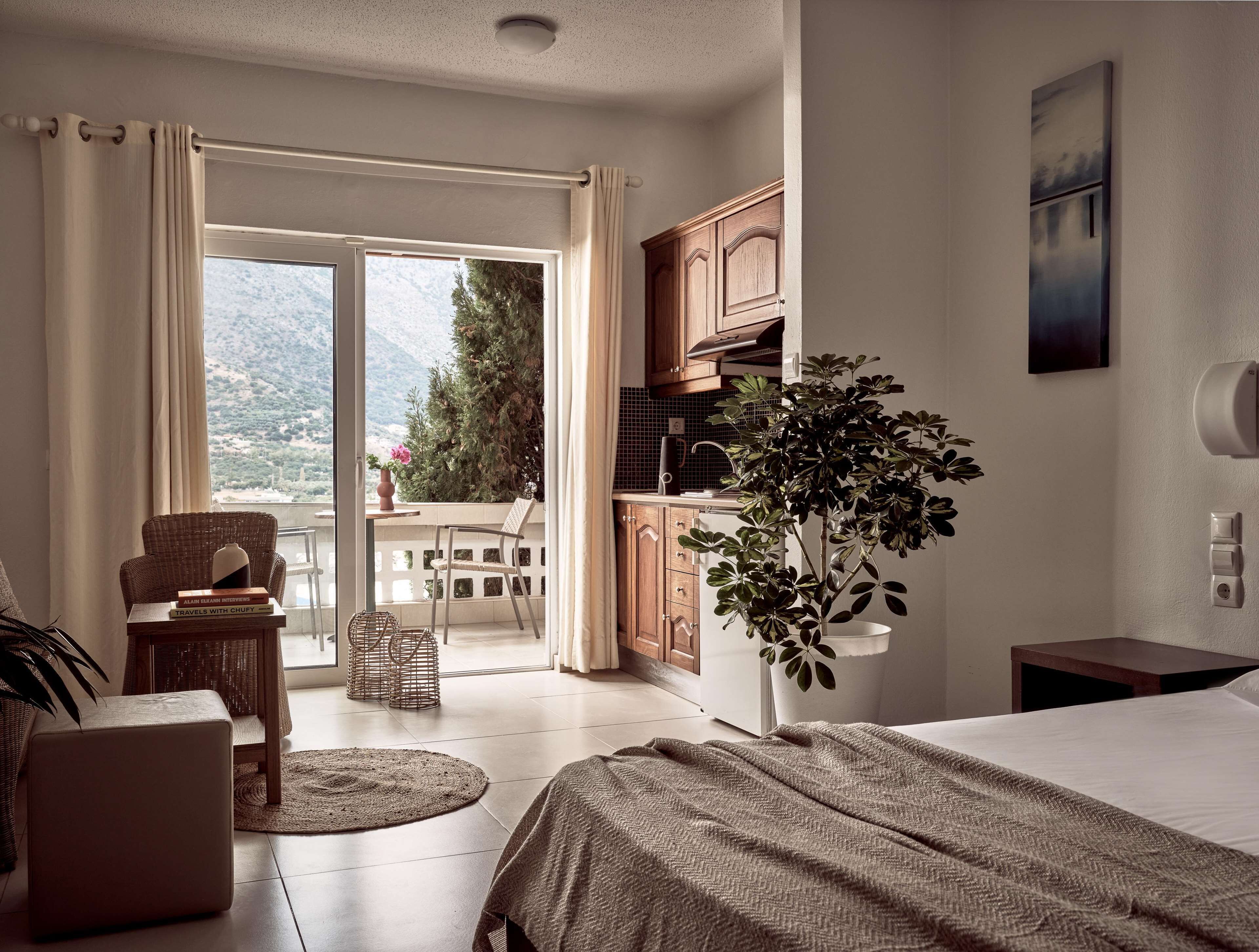 Habitación de hotel con cama, una mesita con libros, una planta en maceta y vistas a un balcón con silla y mesa.