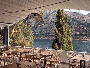 An outdoor seating area with wooden tables and chairs, overlooking a body of water and mountains. - Troulis Aparthotel (Mylopotamos)