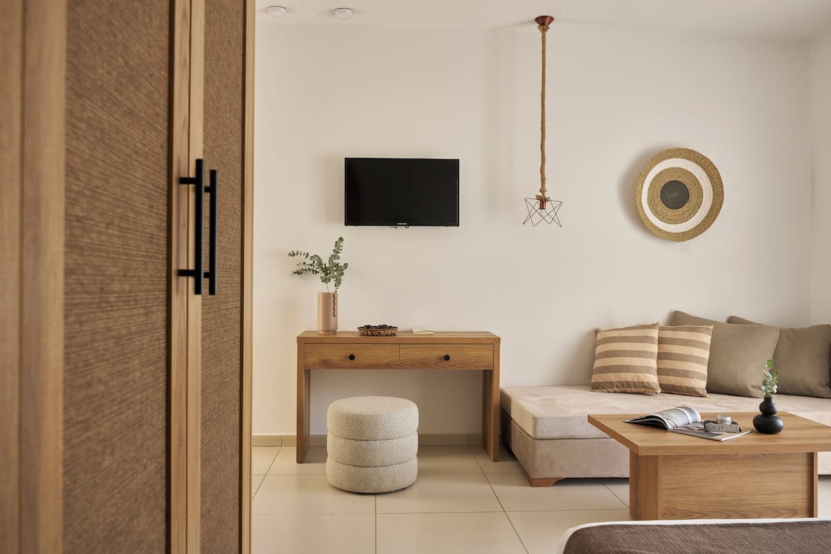 A modern living room with a wooden desk, a round ottoman, a sofa with striped cushions, a coffee table with a magazine and a camera, and a flat-screen TV on the wall.
