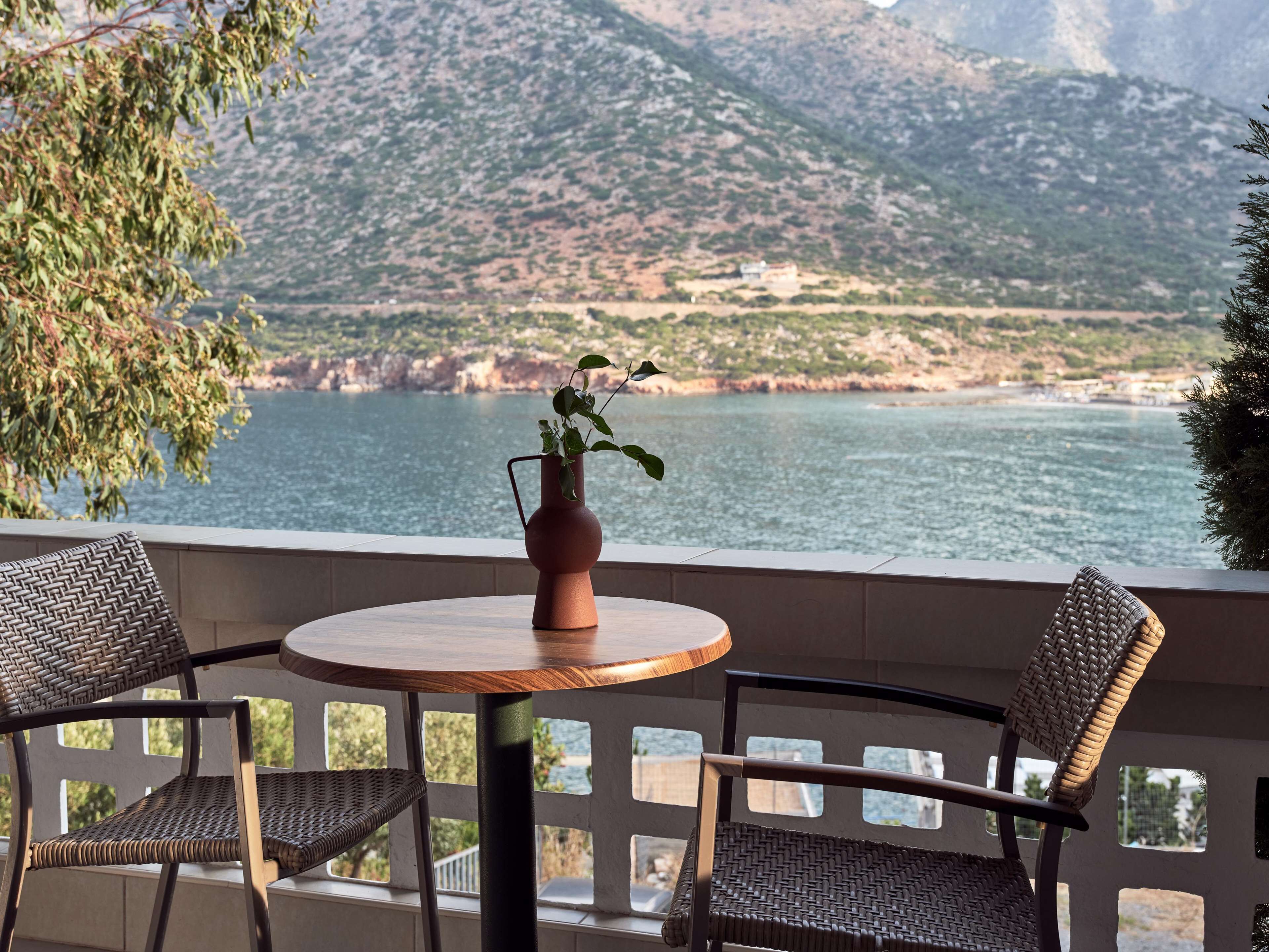 A terrace with a round table and two wicker chairs, overlooking a coastal landscape with mountains and a body of water.