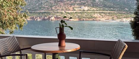 A terrace with a round table and two wicker chairs, overlooking a coastal landscape with mountains and a body of water.