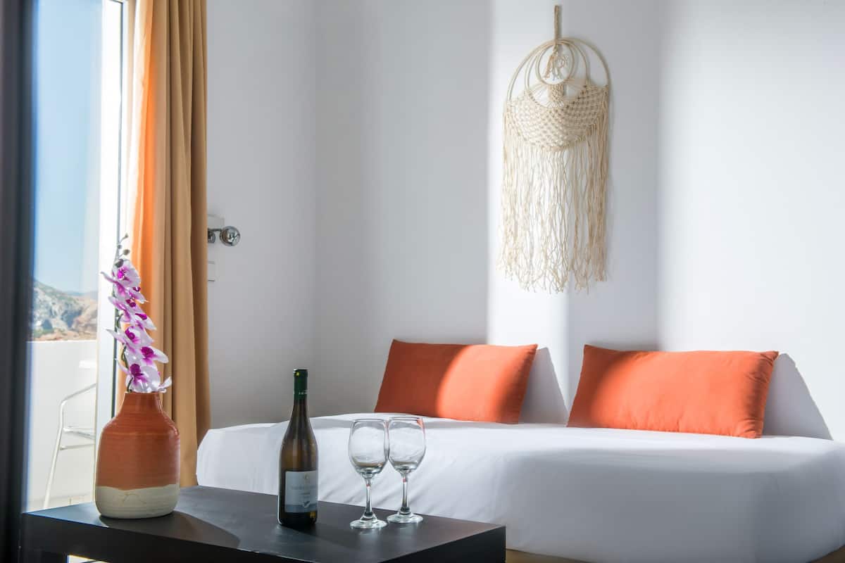 A hotel room with a bed, orange pillows, a vase with flowers, and two wine glasses on a table.