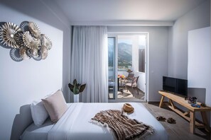 A modern hotel room with a bed, a TV, a balcony with a table and chairs, and a view of mountains. - Troulis Aparthotel (Mylopotamos)