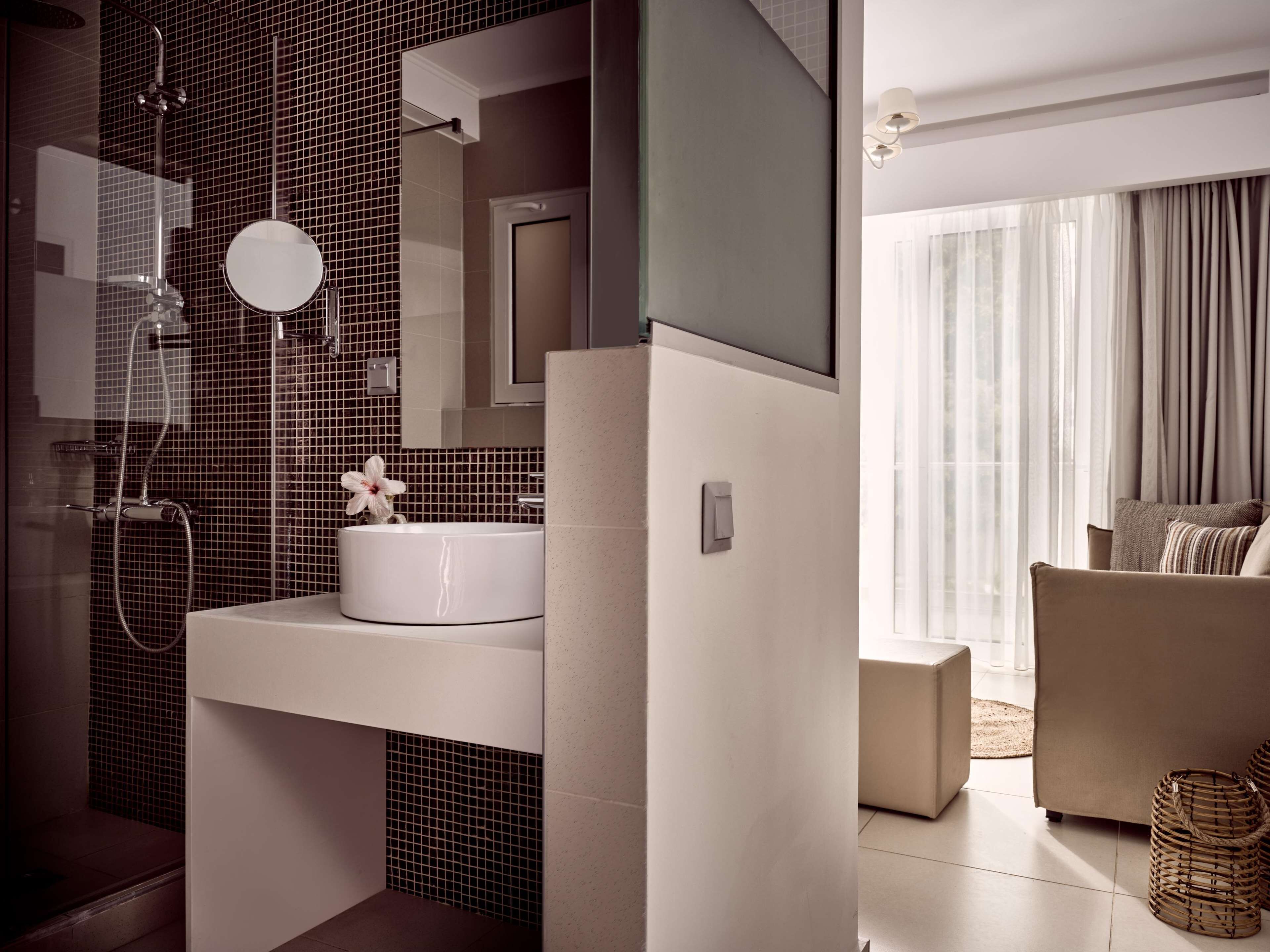 Un baño moderno con un lavamanos blanco, un espejo y una ducha. En la zona de estar contigua, hay un sofá color beige y una ventana con cortinas.