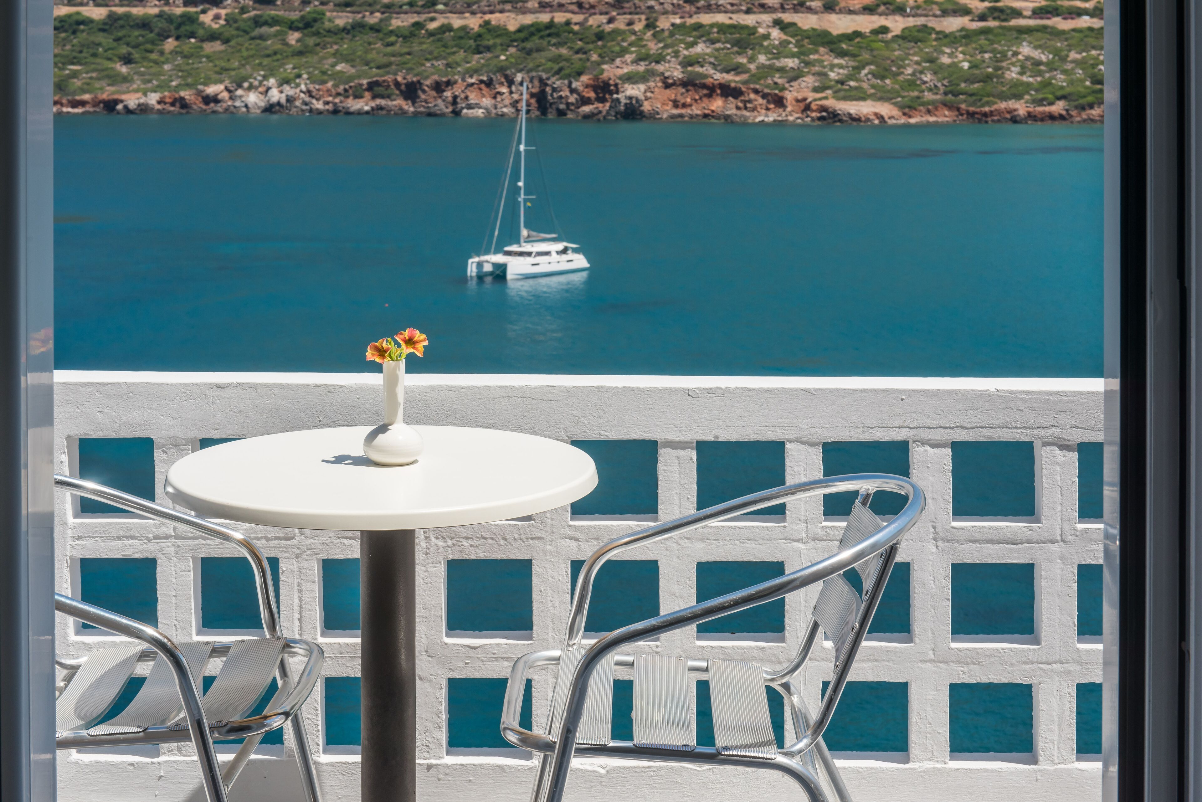 A balcony with a white table and chairs, overlooking a calm sea with a sailboat.