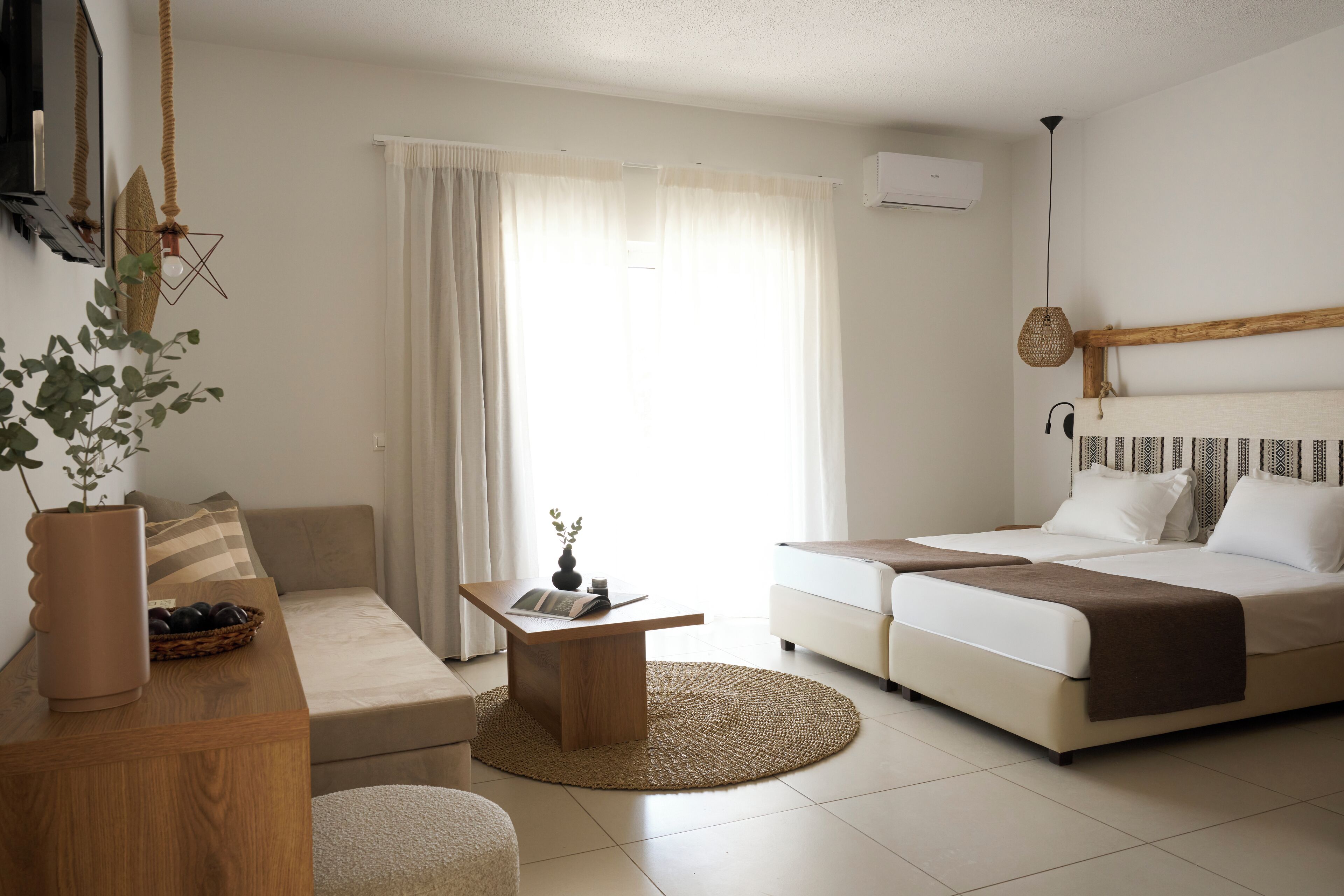 A modern hotel room with a sofa, a small round table, a bed with a headboard, and a large window with curtains.