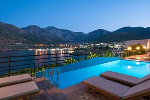 Una piscina infinita con vistas a un lago y montañas, sillas de descanso y una barandilla.