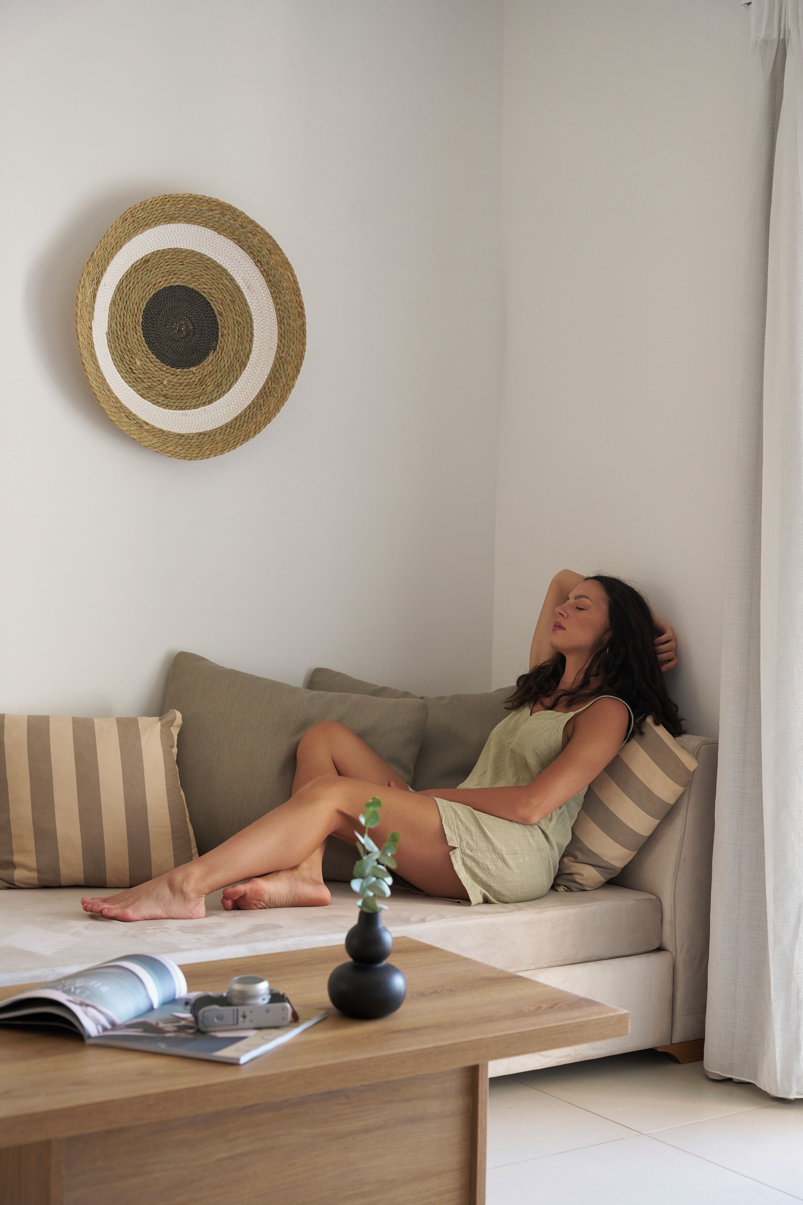 A person reclining on a sofa with striped pillows, a hat on the wall, and a magazine on a wooden table.
