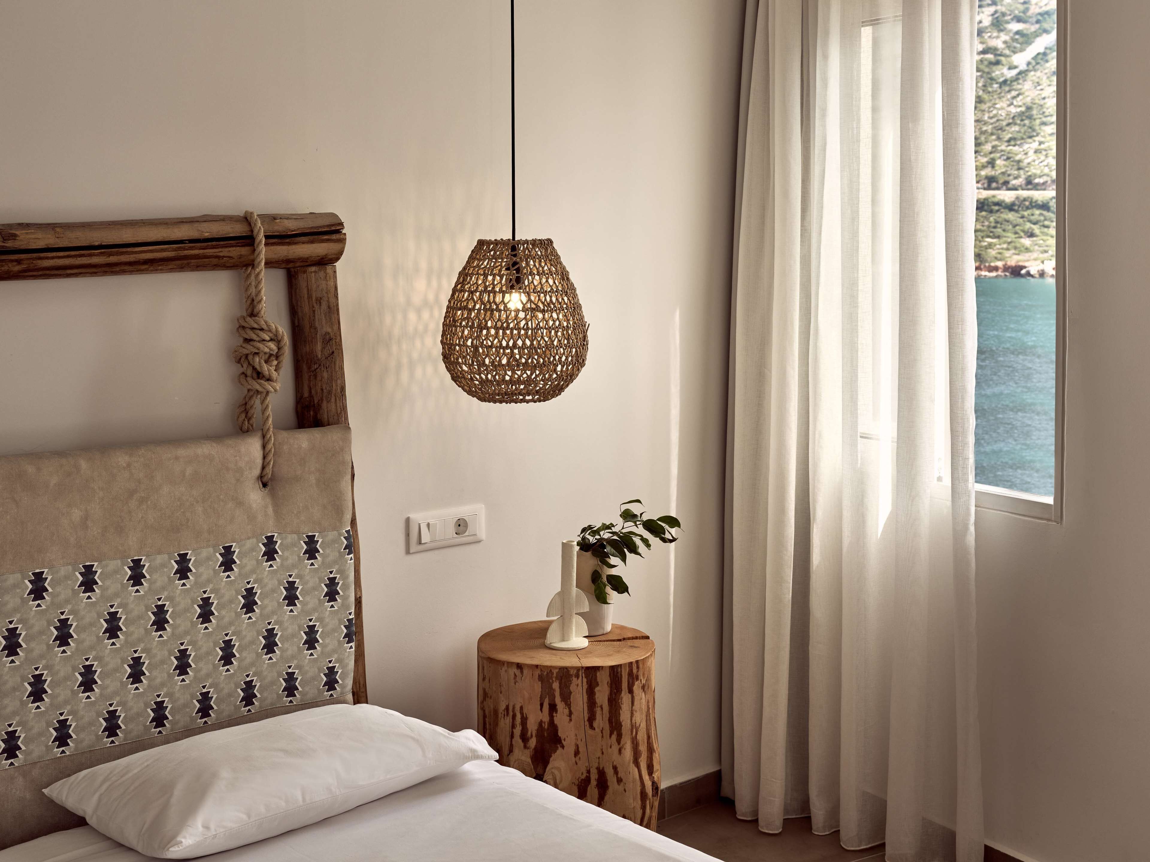 A bedroom with a bed, a bedside table with a plant, a hanging woven light fixture, and a view of the sea through sheer curtains.
