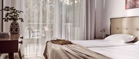 A hotel room with a bed, a desk with a vase, and a view of the outdoors through sheer curtains.