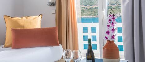 Habitación de hotel con cama, dos almohadas, una mesa con una botella y dos vasos, y vista a un edificio desde la ventana.