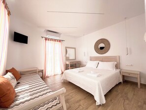 A hotel room with a bed, a bedside table, a chair, a TV, a mirror, and a window with curtains. - Cyclades Blue (Mykonos)
