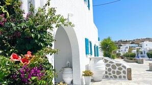 A white building with blue shutters, potted plants, and a stone-paved pathway. - Cyclades Blue (Mykonos)