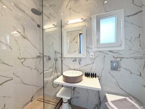 Un baño moderno con ducha acristalada, pared con azulejos de mármol, lavamanos central con un lavabo redondo y una pequeña ventana sobre el espejo.