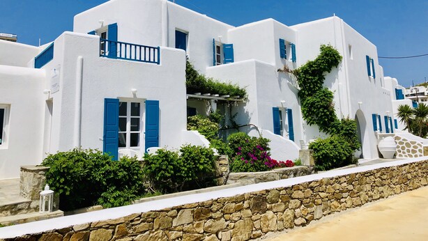 A white building with blue shutters and a stone wall in front. - Cyclades Blue (Mykonos)