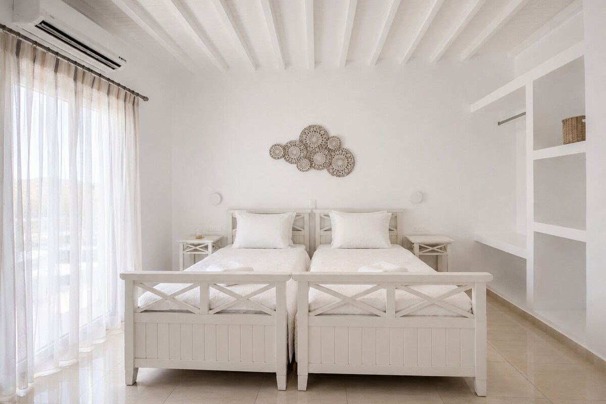 A bedroom with two beds, a ceiling fan, a wall-mounted air conditioner, and a large window with sheer curtains.