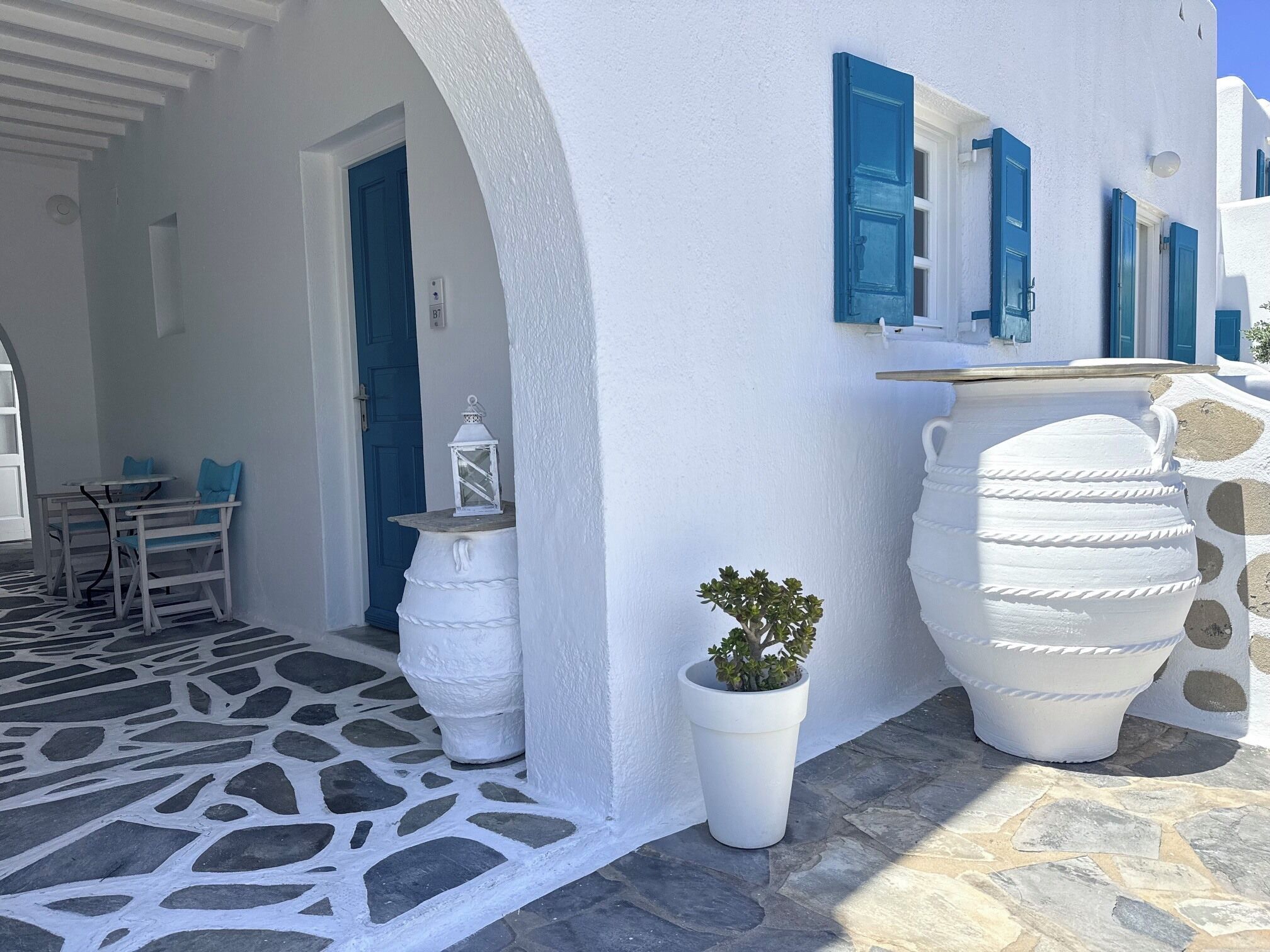 A white building with blue shutters, a stone floor, and a potted plant.