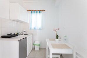 A small, clean kitchen with white cabinets, a dishwasher, a sink, and a window with blue curtains. There is a small table with a potted plant and two water bottles. - Cyclades Blue (Mykonos)