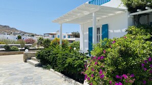 A white building with blue shutters and a covered porch, surrounded by lush greenery and purple flowers. - Cyclades Blue (Mykonos)