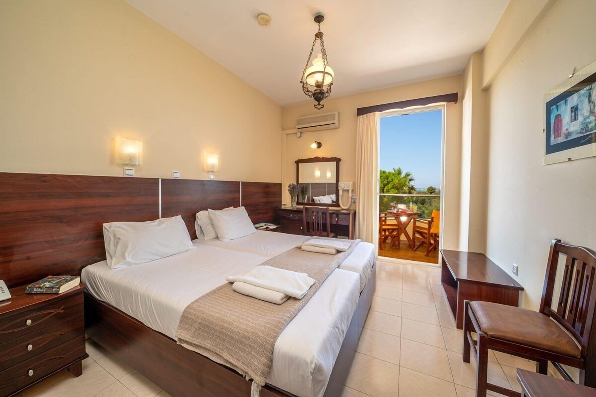 A hotel room with a large bed, a wooden headboard, a desk with a chair, a balcony with a table and chairs, and a hanging light fixture.