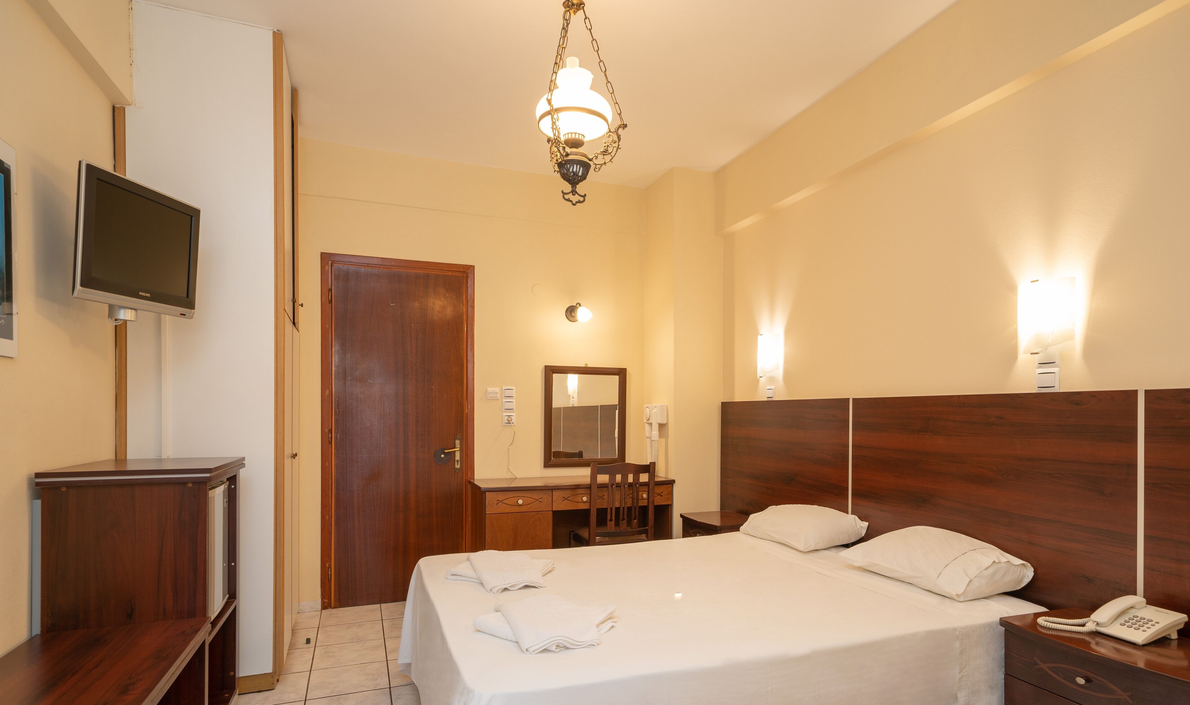 Una habitación de hotel con cama, mesita de noche, lámpara, espejo y televisor sobre un soporte.