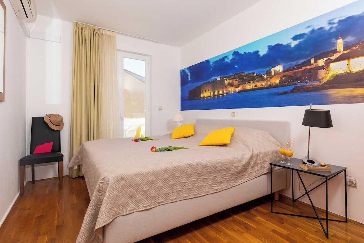 A bedroom with a large bed, a chair, a nightstand, and a painting of a cityscape.