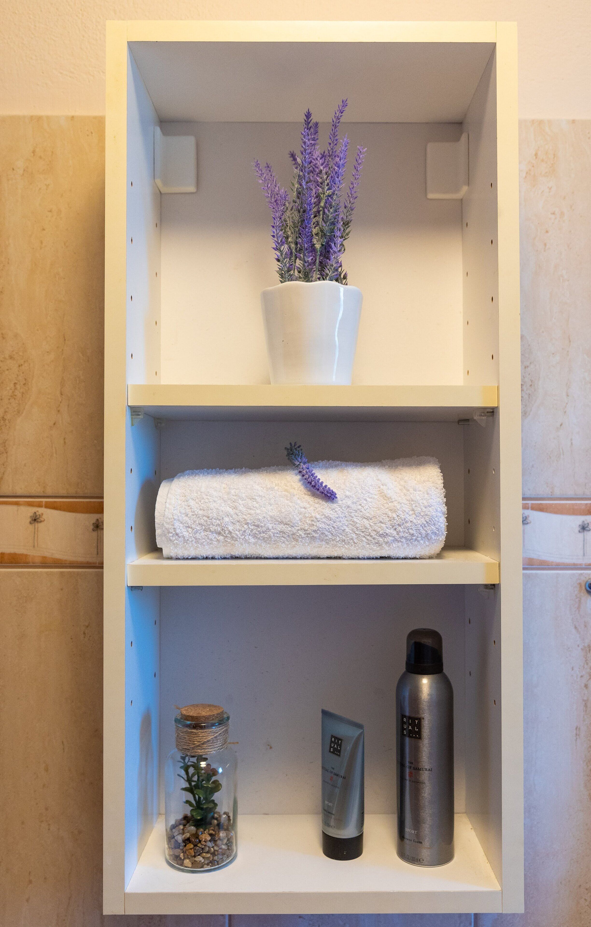 Una estantería con una planta de lavanda, una toalla doblada, un frasco pequeño con una planta y dos botellas de artículos de aseo.