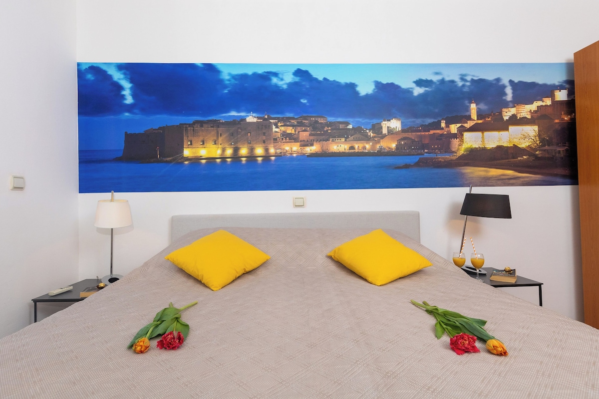 A hotel room with a bed, two nightstands, a lamp, and a large cityscape painting on the wall.