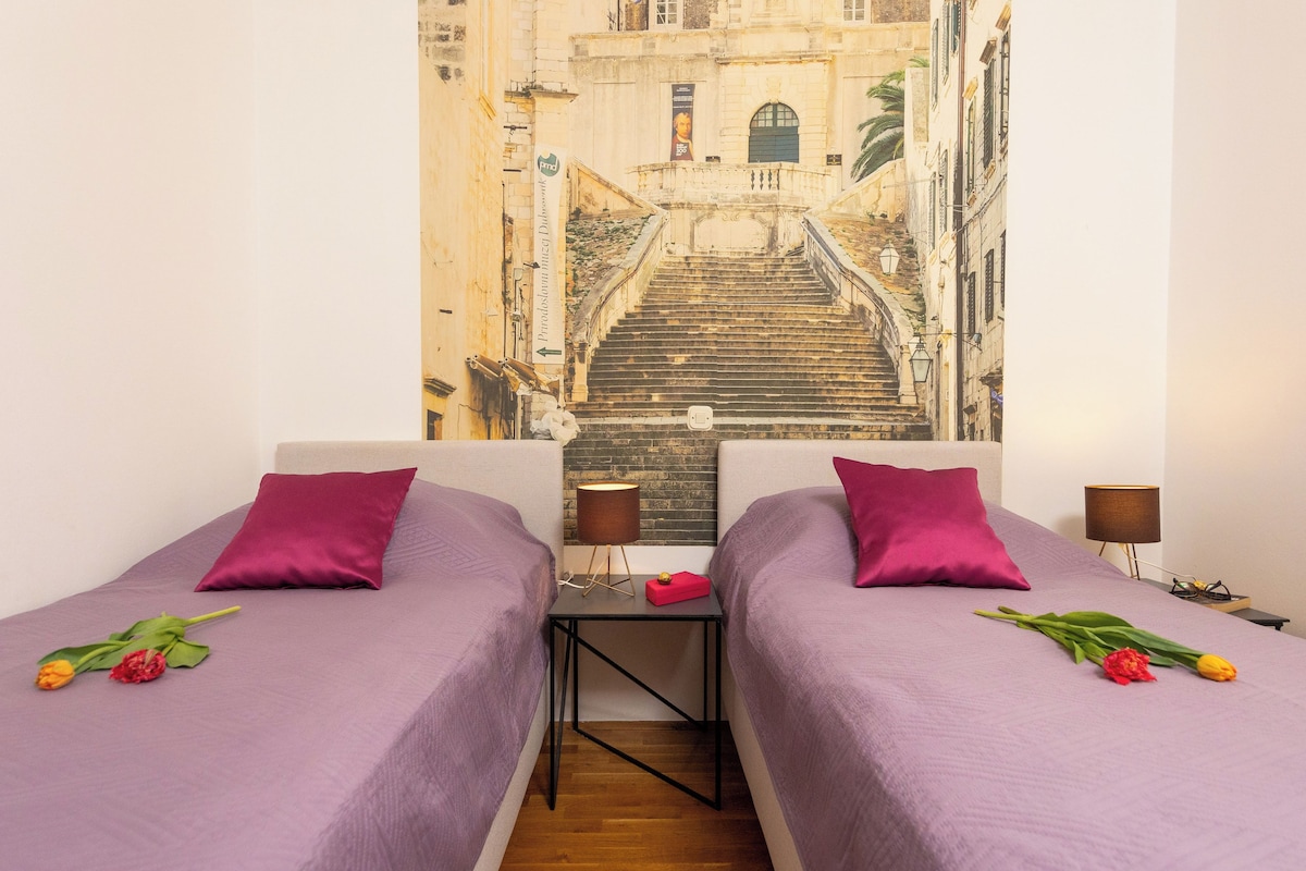A room with two beds, each with a purple bedspread and a red pillow. There is a small table between the beds with a lamp and a red box. A painting of a street scene is on the wall behind the beds.