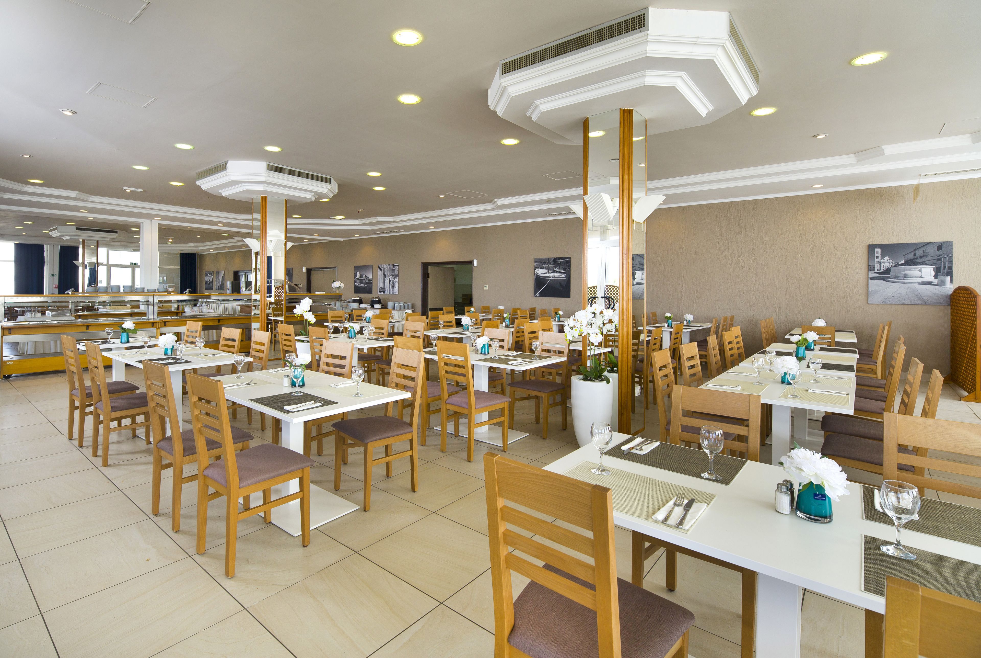 Un grand espace dédié à la salle à manger, avec plusieurs tables et chaises, des bouches d’aération pour la climatisation et des œuvres d’art encadrées accrochées aux murs.