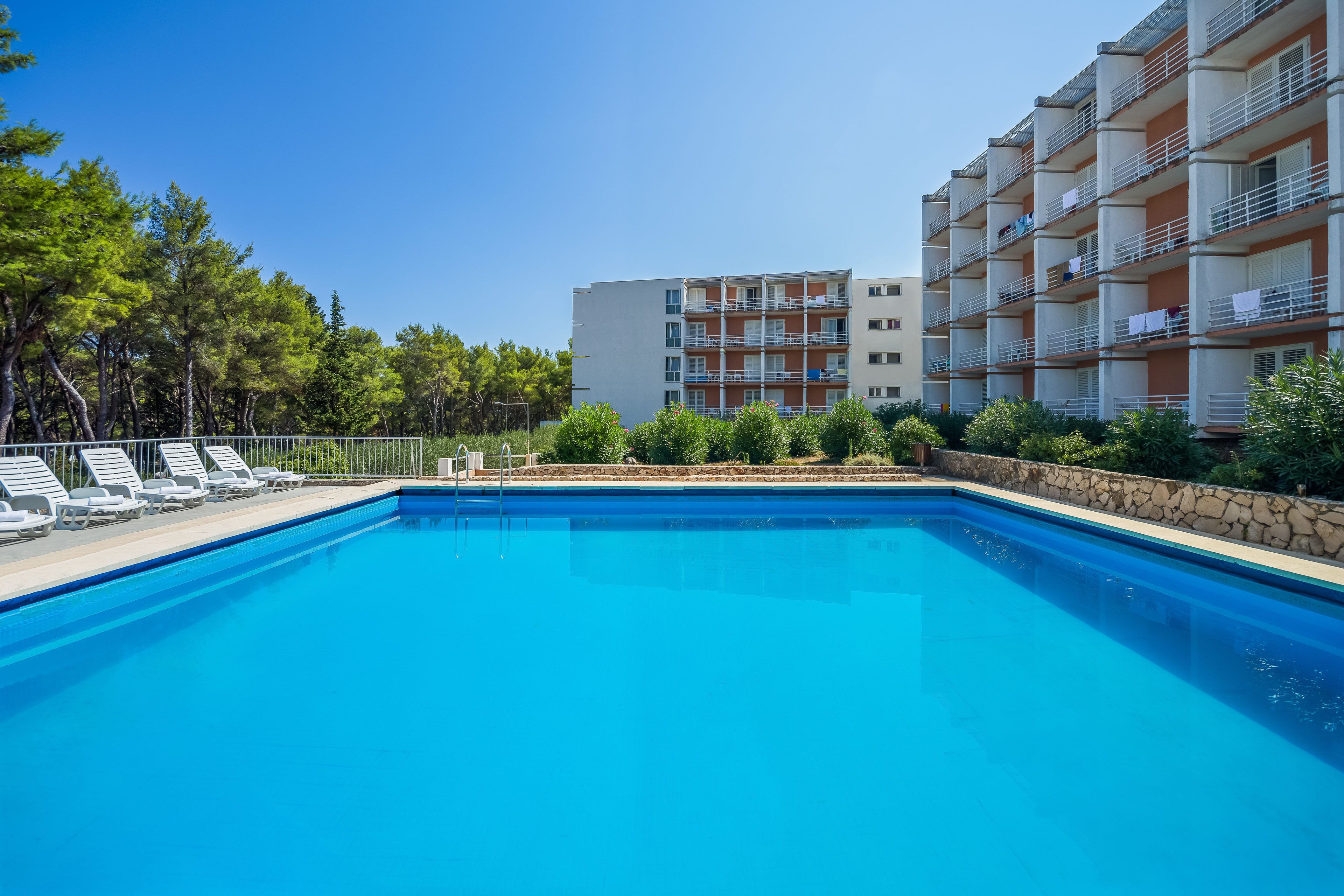 A swimming pool with lounge chairs, a stone wall, and a multi-story building with balconies.
