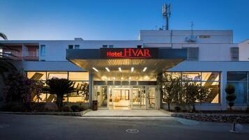 A modern hotel entrance with a clear sky, a sign reading "Hotel HVAR," and a well-lit facade.