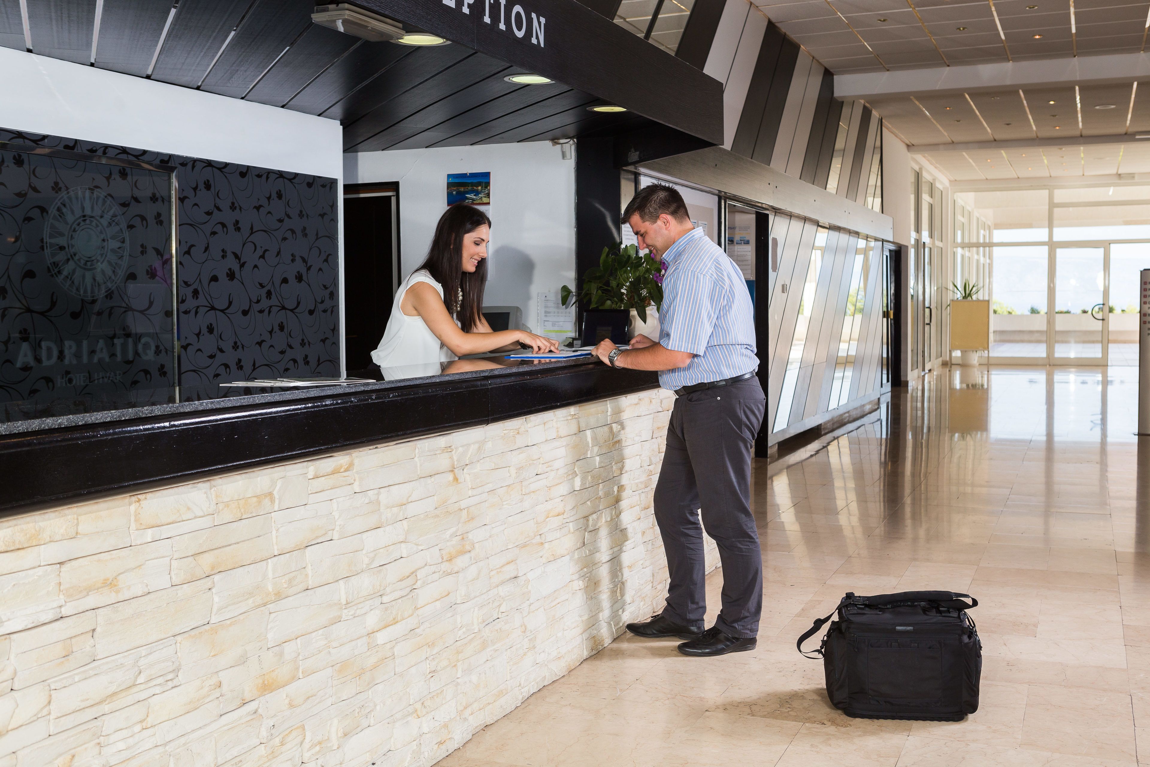 A reception area with a black counter, a receptionist, and a man standing. There is a black bag on the floor.