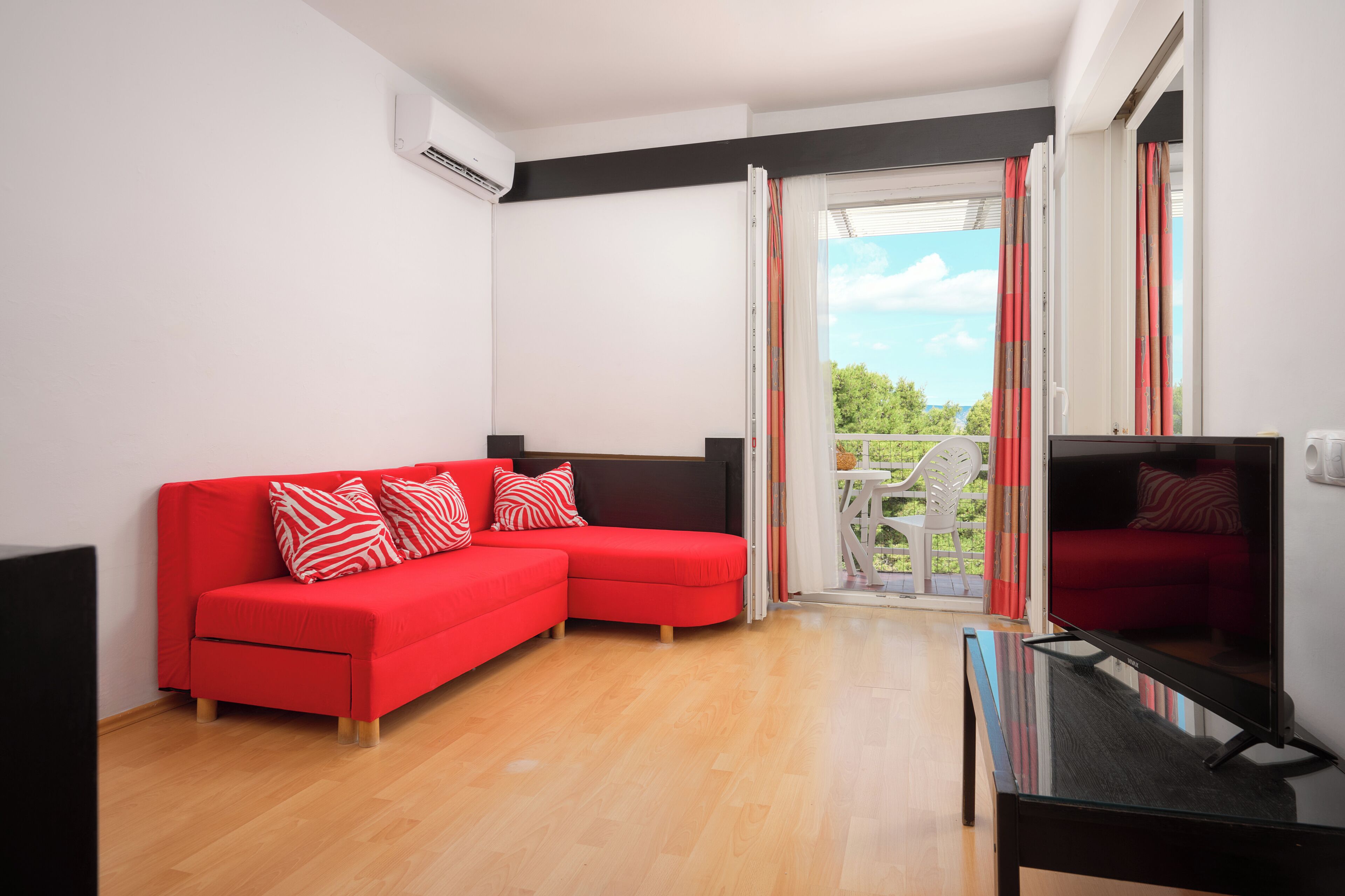 Un salon moderne avec un canapé d’angle rouge, un téléviseur à écran plat et un balcon offrant une vue sur la verdure.