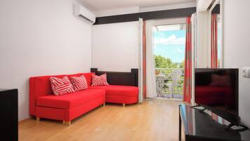 Un salon moderne avec un canapé d’angle rouge, un téléviseur à écran plat et un balcon offrant une vue sur la verdure.