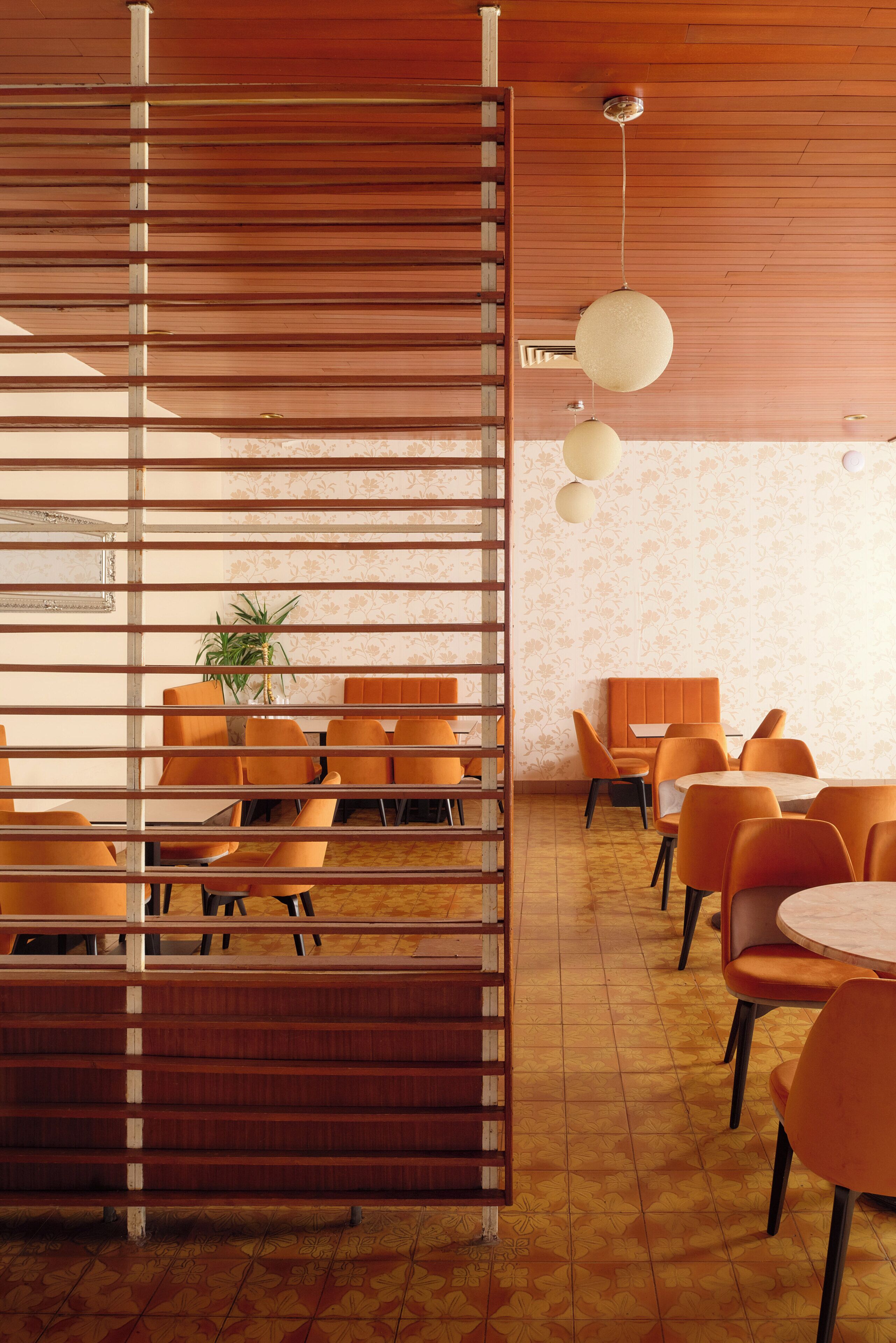 Un comedor moderno con sillas y mesas anaranjadas, un separador de madera y un muro con diseño.