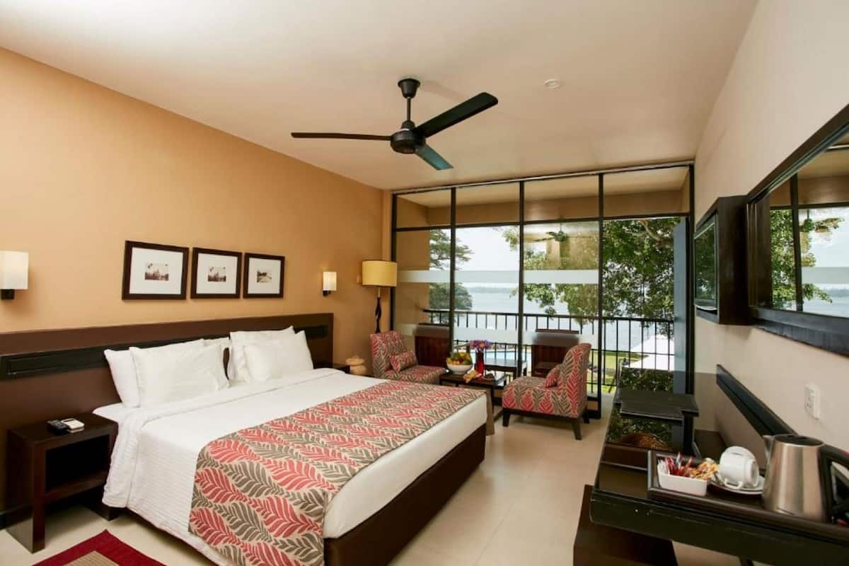 A hotel room with a bed, two chairs, a desk, a mirror, a ceiling fan, and a balcony with a view.