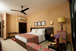 A hotel room with a bed, a ceiling fan, an air conditioning unit, a nightstand, a lamp, a chair, and a small table with a vase of flowers. - Ekho Safari Tissa Hotel (Thissamaharama)