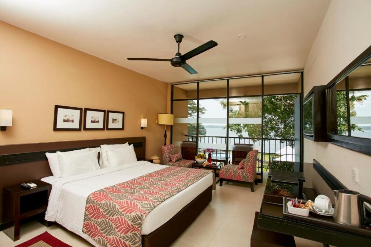 A hotel room with a bed, two chairs, a desk, a mirror, a ceiling fan, and a balcony with a view.