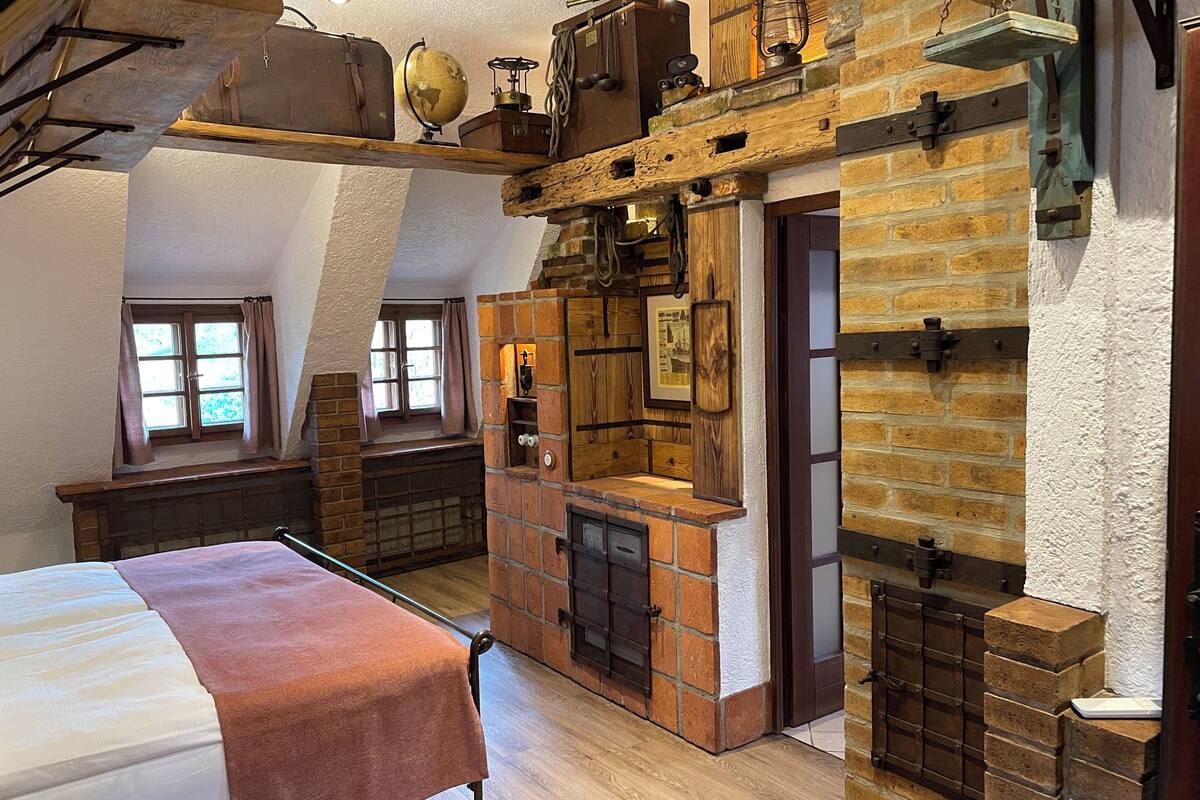 A loft-style room with a bed, a wooden desk, a brick fireplace, and a window with curtains.