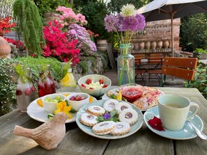A table set outdoors with a variety of food items, including pastries, fruits, and drinks, with a garden and brick wall in the background.