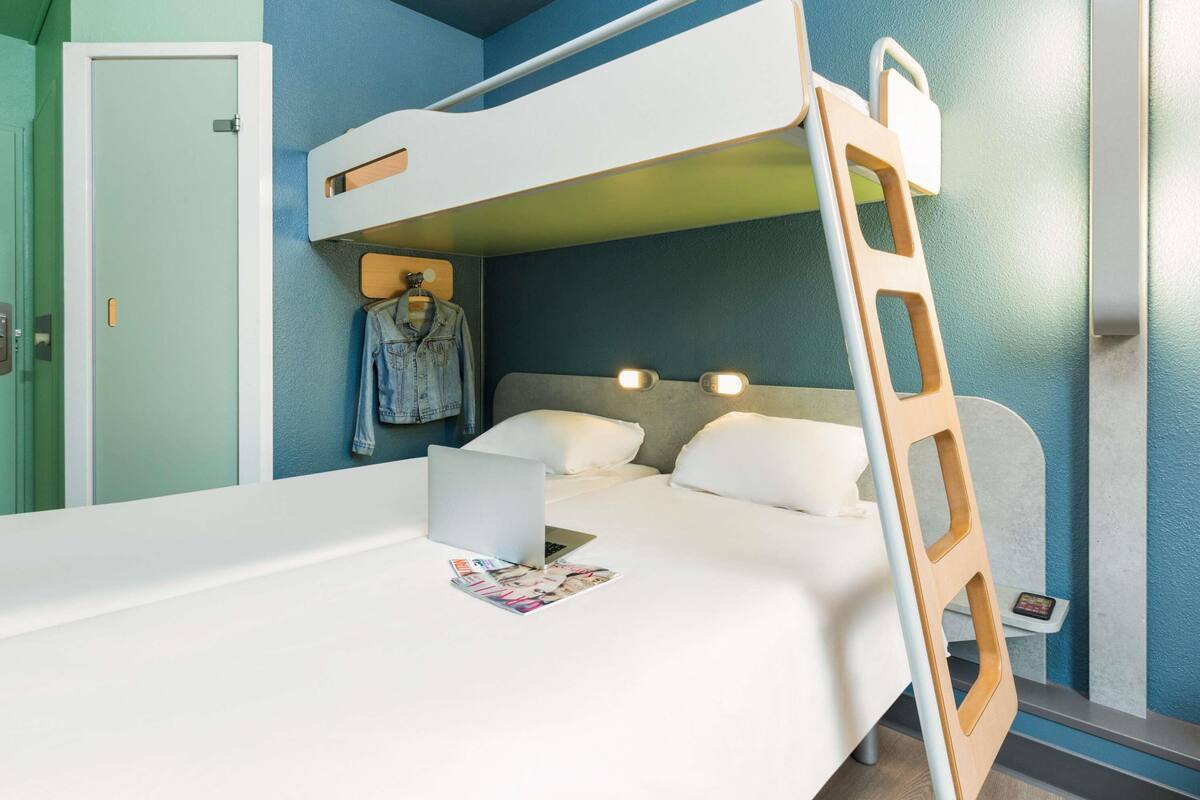 Habitación con literas, una computadora portátil y revistas sobre la cama, una escalera de madera y una chaqueta vaquera colgada en la pared.