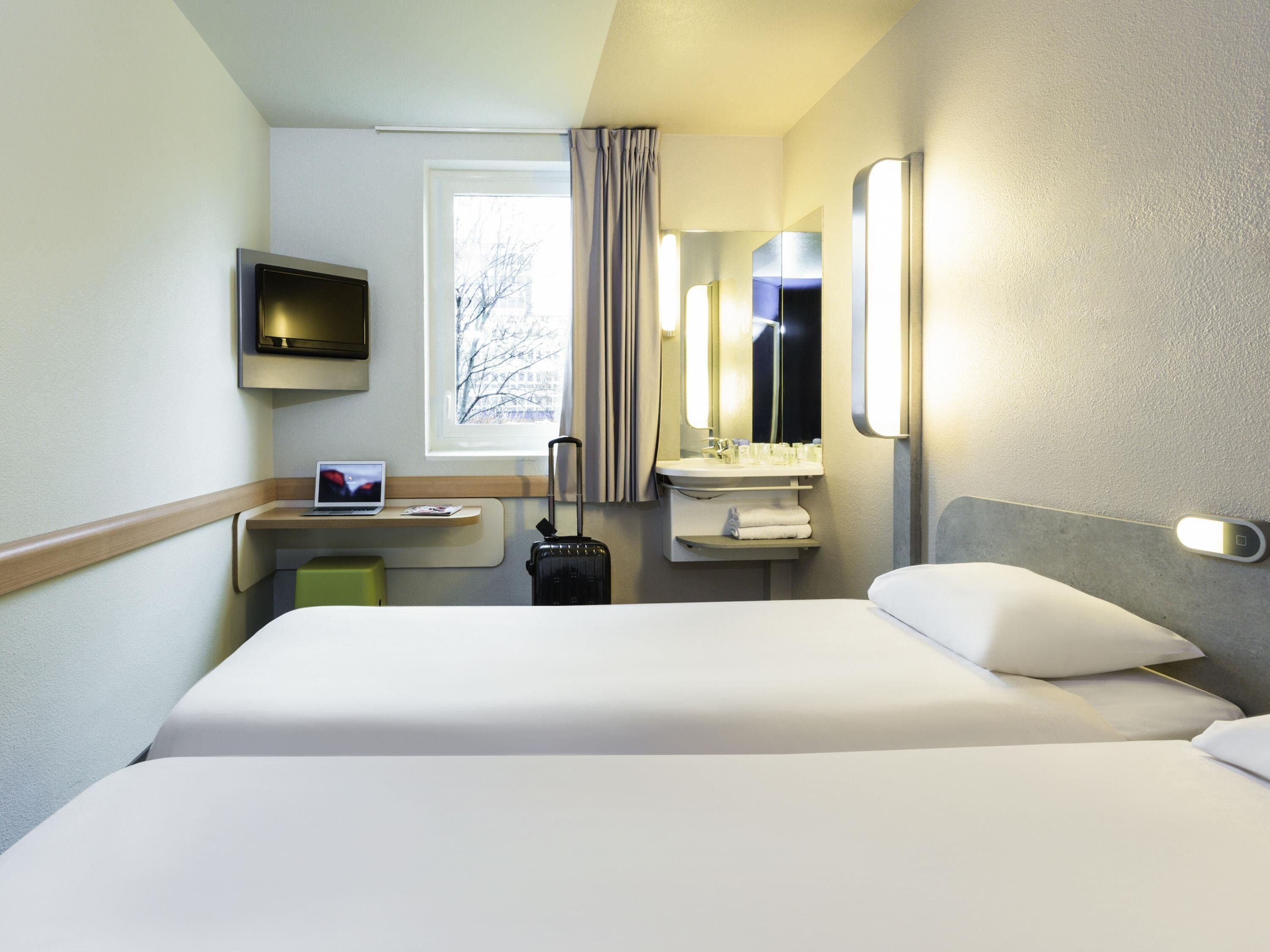 A hotel room with a bed, a desk with a laptop, a suitcase, a TV, a window with curtains, and a bathroom with a mirror and sink.