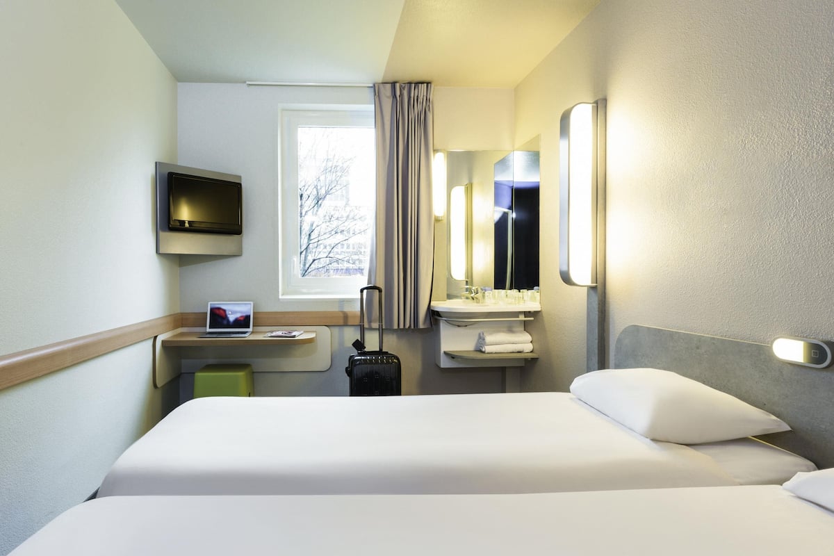 A hotel room with a bed, a desk with a laptop, a suitcase, a TV, a window with curtains, and a bathroom with a mirror and sink.
