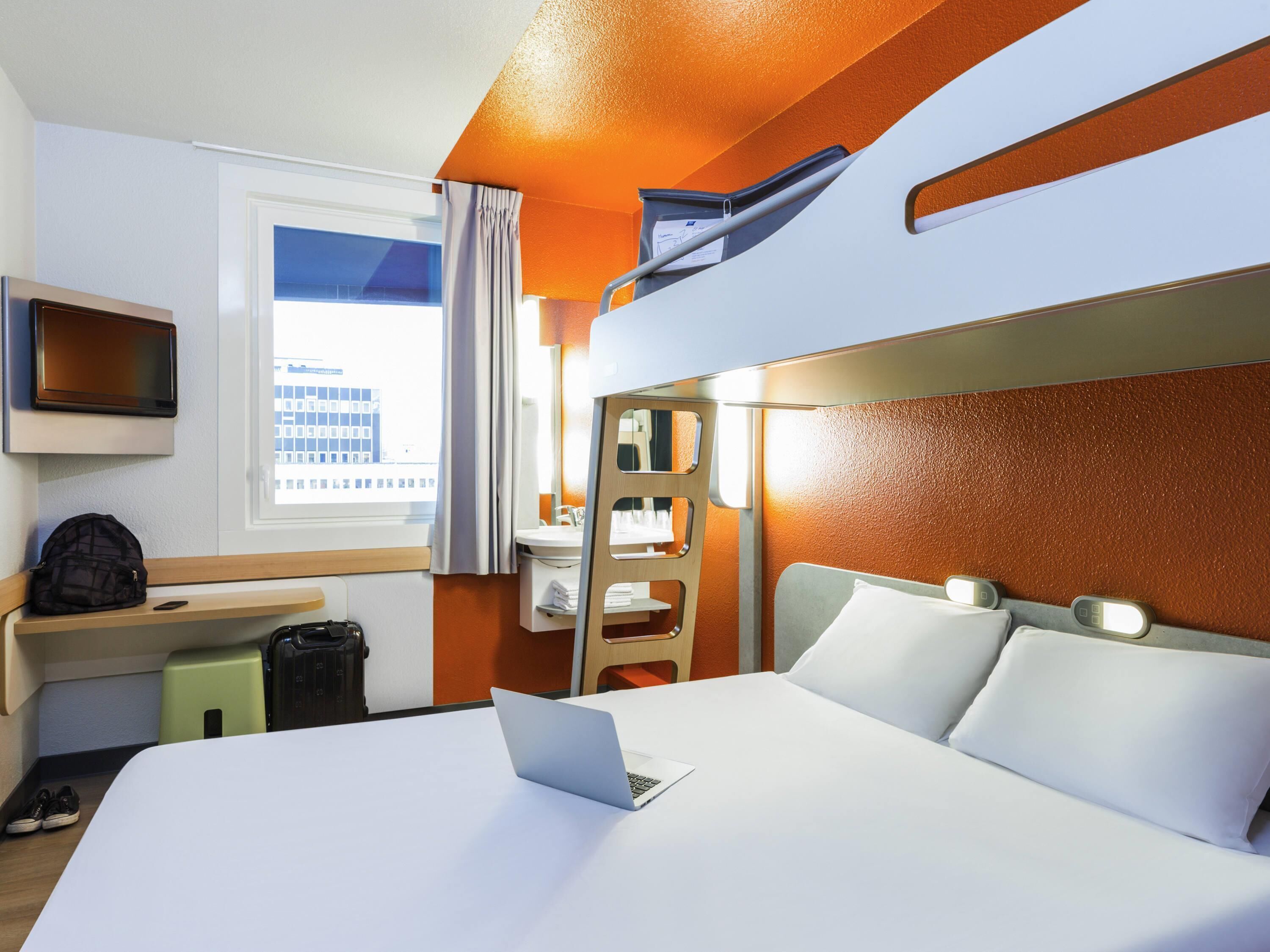A hotel room with a bed, a desk with a laptop, a TV, and a window with curtains.