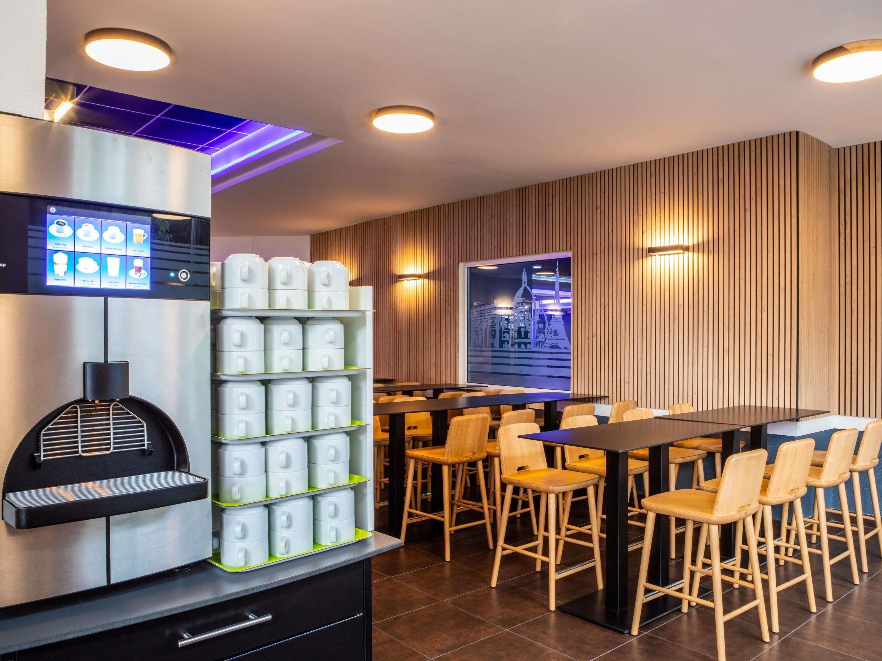 Un espace bar moderne, doté d’une machine à café, de piles de tasses et d’un coin salon avec des chaises et des tables en bois.
