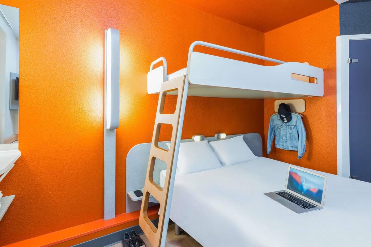 Una cama litera con una computadora portátil en la cama inferior, una escalera de madera y un maniquí con una chaqueta vaquera.