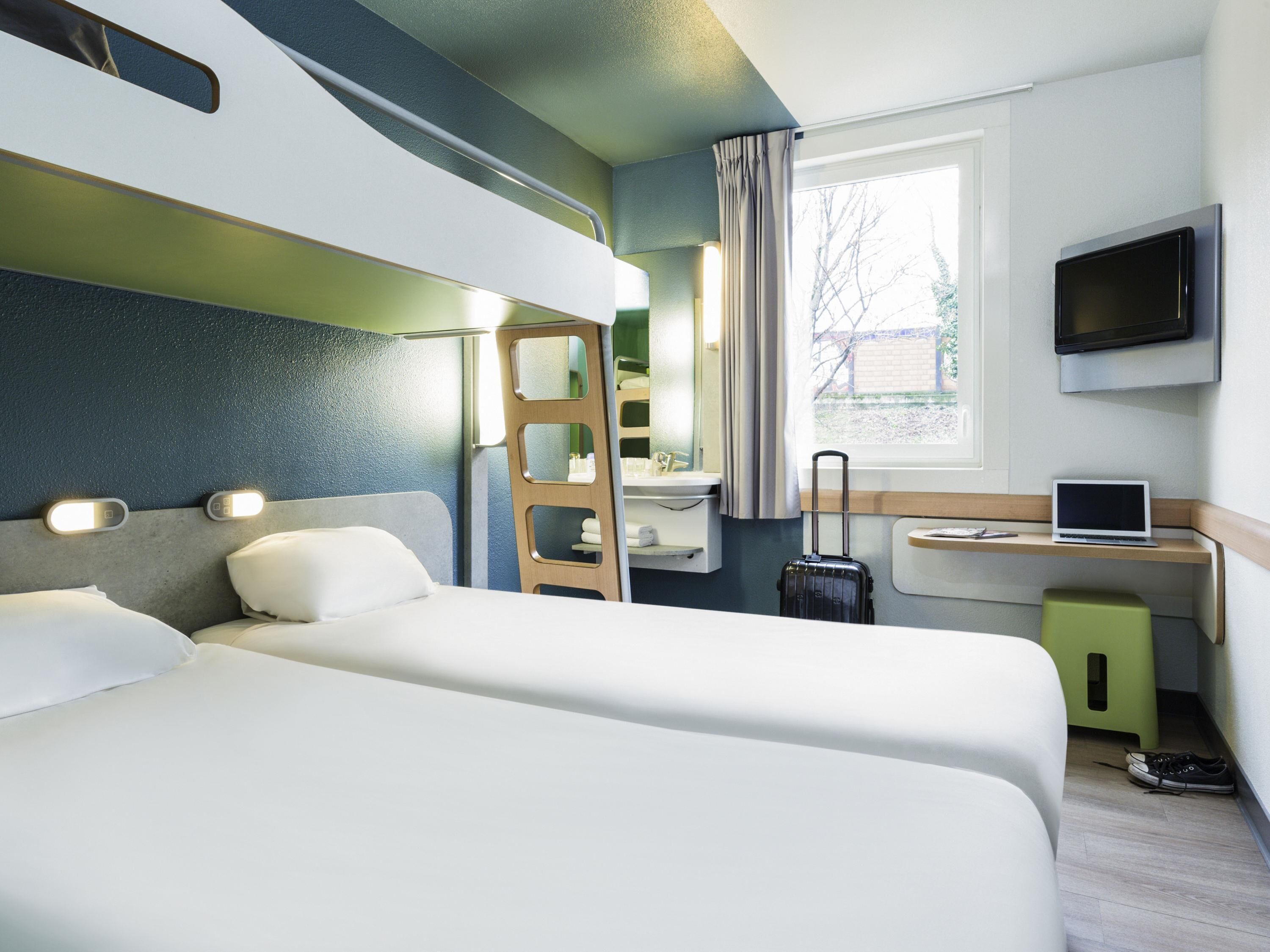 Una habitación de hotel con dos camas, un escritorio con una computadora portátil, un televisor, una maleta y una ventana con cortinas.