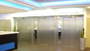 Un área de ascensores moderna con puertas metálicas, un mostrador de recepción y plantas en macetas.