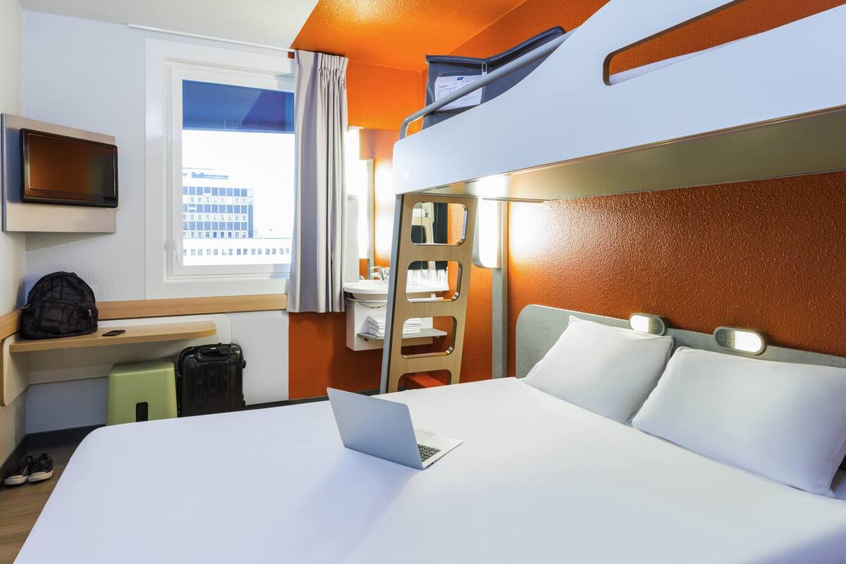 A hotel room with a bed, a desk with a laptop, a TV, and a window with curtains.